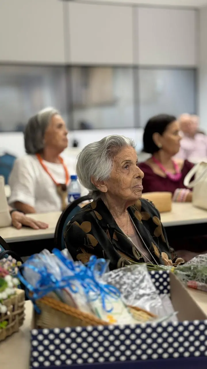 Na imagem, uma fotografia de média distância, com foco raso, mostra uma mulher idosa de cabelos curtos e grisalhos, a Dona Maria Augusta, sentada numa mesa no primeiro plano. Ela veste um blazer preto com padrão floral em tons de cobre e um colar prateado fino. O seu olhar está virado para a direita da imagem, para fora de campo. À sua frente, no primeiro plano, há uma caixa de presentes aberta com base azul escura às bolinhas brancas, contendo lembranças embaladas com fitas azuis e um pequeno cesto de verga. Duas outras mulheres e um homem estão sentados em mesas atrás dela, desfocados mas visíveis. O fundo é um ambiente de sala de aula ou escritório.