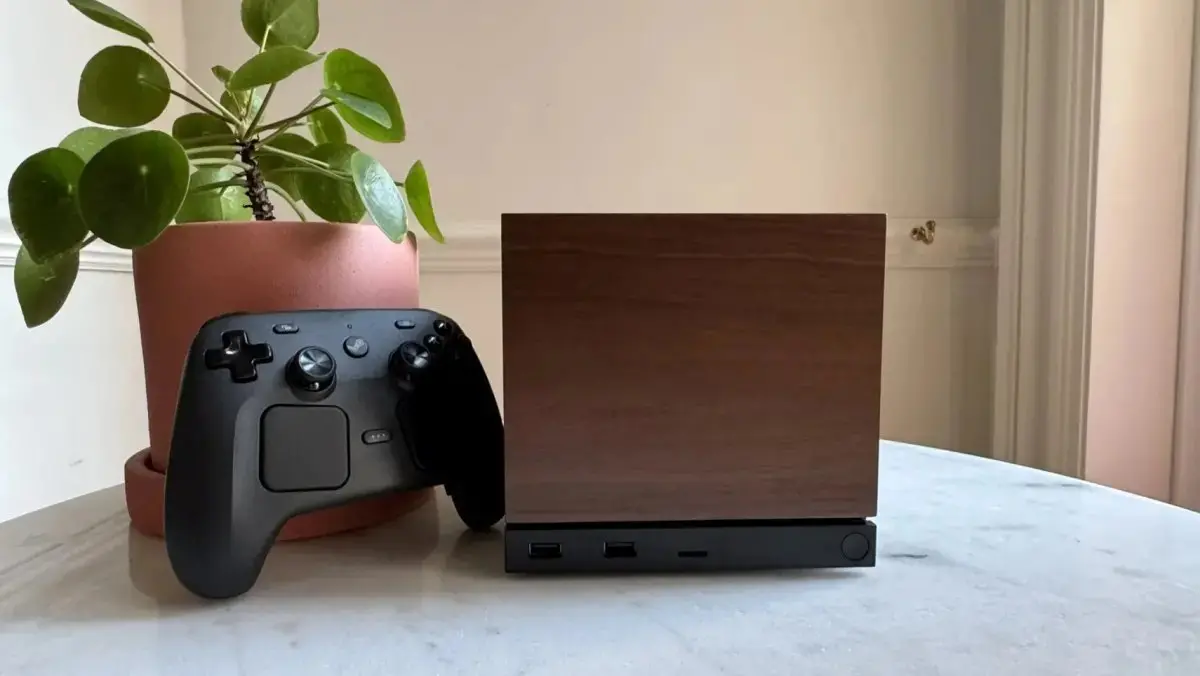 Conjunto com controle de videogame preto apoiado ao lado de uma caixa/console de madeira escura sobre uma mesa clara, com planta em vaso ao fundo.