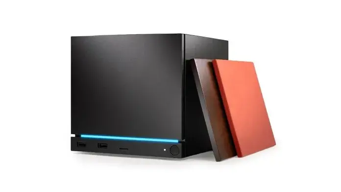 Conjunto com controle de videogame preto apoiado ao lado de uma caixa/console de madeira escura sobre uma mesa clara, com planta em vaso ao fundo.