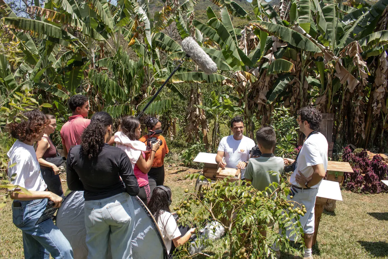 Uma equipe de filmagem jovem e com câmeras grava um homem ao lado de caixas de colmeias em um jardim gramado, cercado por um denso bananal ao fundo. Na cena, um microfone de haste (boom) é estendido sobre o grupo enquanto o homem sorri e interage com os participantes.