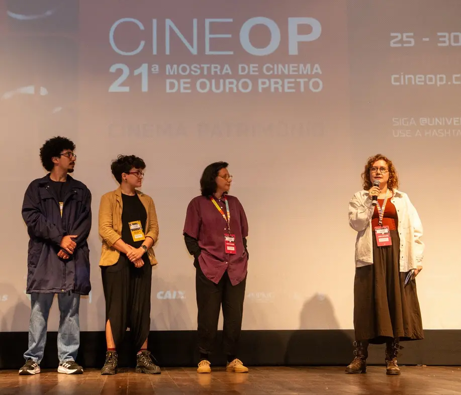 Três profissionais da equipe do MIS Ceará estão alinhados no palco do festival CineOP, sendo um homem e duas mulheres. À direita, outra mulher está falando ao microfone enquanto os outros três escutam atentamente. Ao fundo, um grande telão exibe a identidade visual da 21ª Mostra de Cinema de Ouro Preto em tons claros, completando a atmosfera solene e cultural do evento.