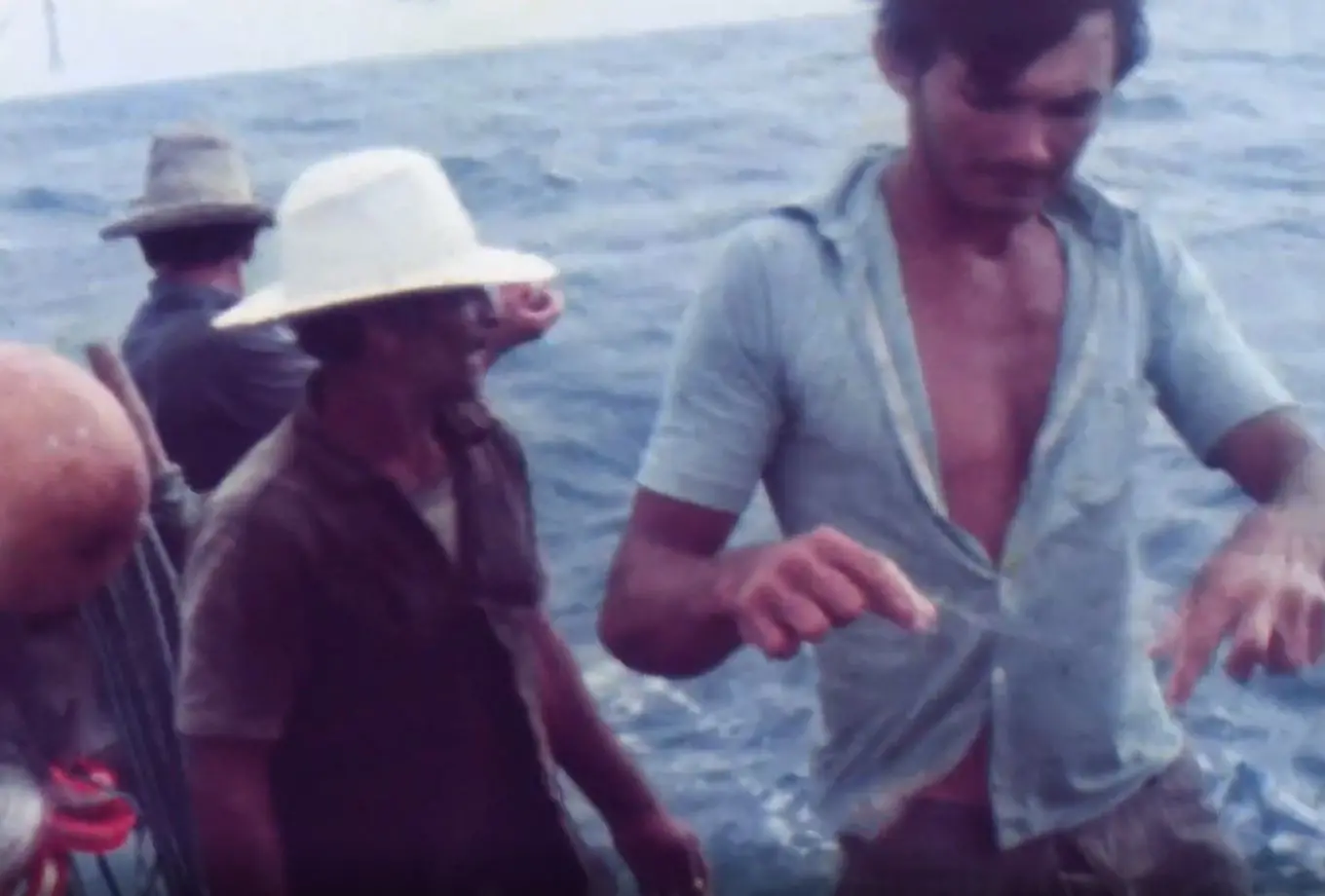 Imagem reproduzida do curta cearense 'Jangada de Ir e Vir' (1977), de Marcus Vale, mostra três pescadores a bordo de uma embarcação em alto-mar, com um homem em primeiro plano à direita manuseando o que parece ser uma linha ou equipamento de pesca. Ao fundo, os outros dois companheiros usam chapéus de palha e observam o oceano azul de águas agitadas, capturados em um registro cinematográfico de aspecto antigo.