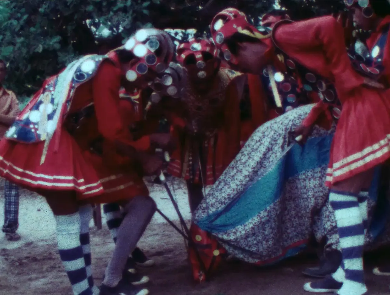 Imagem reproduzida do curta cearense 'Reis do Cariri', de Oswald Barroso, que mostra integrantes de reisado vestindo trajes vermelhos ornamentados e chapéus decorados com espelhos. Eles se inclinam em círculo durante uma apresentação tradicional. Eles utilizam espadas e interagem com uma figura no chão, capturados em um registro com textura cinematográfica antiga.