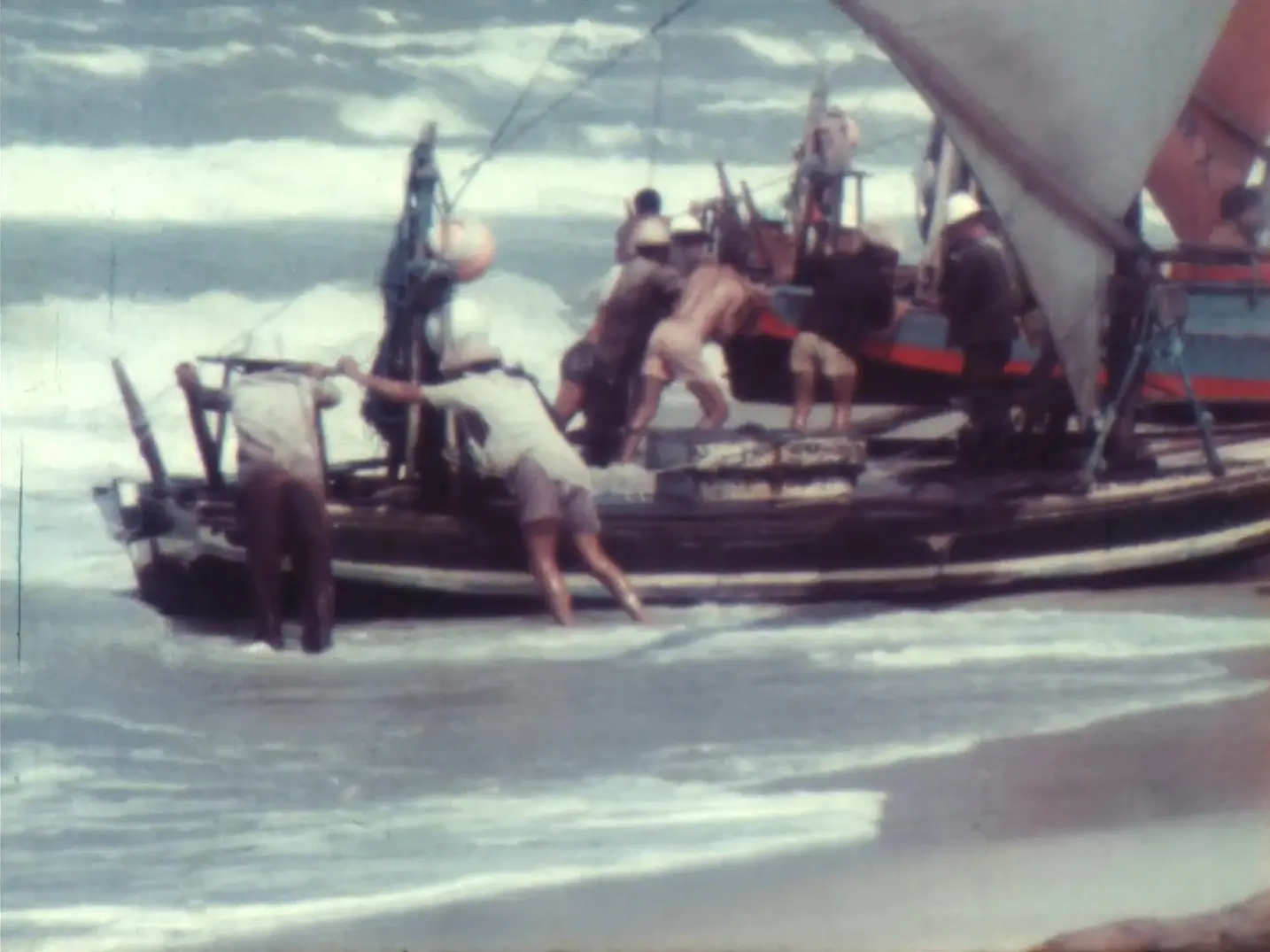 Imagem reproduzida do curta cearense 'Jangada de Ir e Vir', de Marcus Valle, que mostra vários pescadores unindo forças para empurrar uma jangada tradicional contra as ondas na beira da praia. A cena, capturada em uma película antiga com tons desbotados, mostra o grupo trabalhando coordenadamente em meio à espuma do mar e sob ondas agitadas ao fundo.