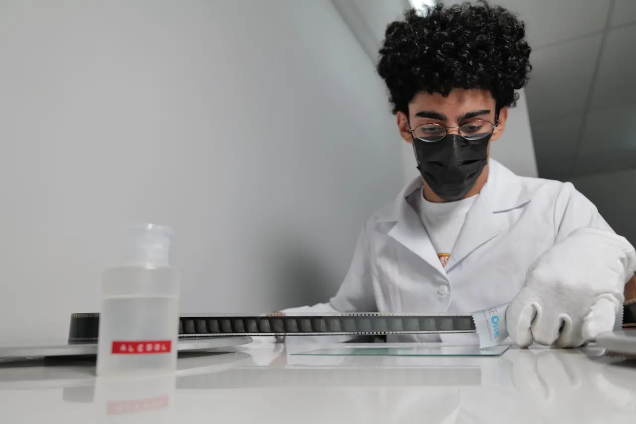 Um profissional de jaleco branco, máscara preta e luvas manuseia cuidadosamente uma película de filme cinematográfico sobre uma bancada branca de laboratório. Ao fundo, o ambiente clean destaca a ação focada do técnico, enquanto em primeiro plano há um frasco com a etiqueta 'ÁLCOOL' desfocado.