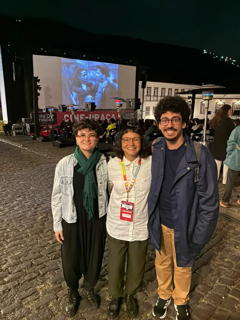 Três pessoas sorriem abraçadas em uma praça de paralelepípedos durante uma exibição de cinema ao ar livre no período noturno. Ao fundo, um grande telão exibe um filme clássico em preto e branco diante de uma plateia sentada, sob a estrutura do evento identificada como 'CINE-PRAÇA'.
