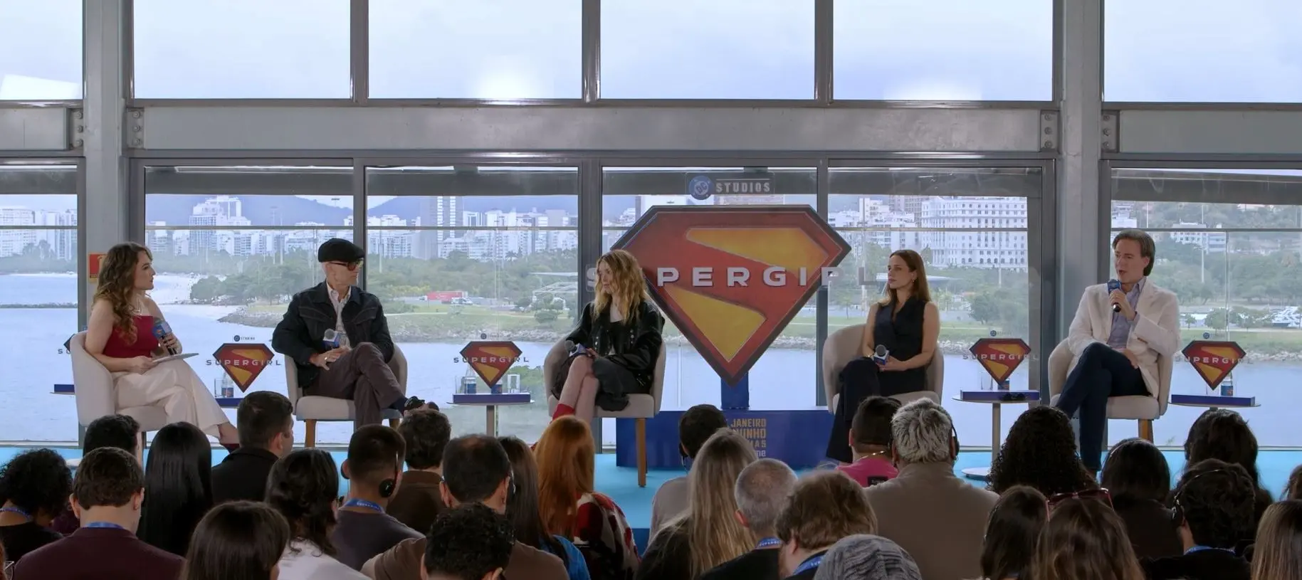 Painel de apresentação realizado em um auditório com público sentado em primeiro plano. Quatro participantes estão acomodados em poltronas sobre um palco, cada um segurando um microfone. Ao centro, há um grande painel com o símbolo da Supergirl e o nome da personagem. Ao fundo, paredes de vidro revelam a vista de uma área urbana à beira d’água, com prédios e vegetação. Mesas laterais com garrafas de água e placas temáticas completam o cenário.