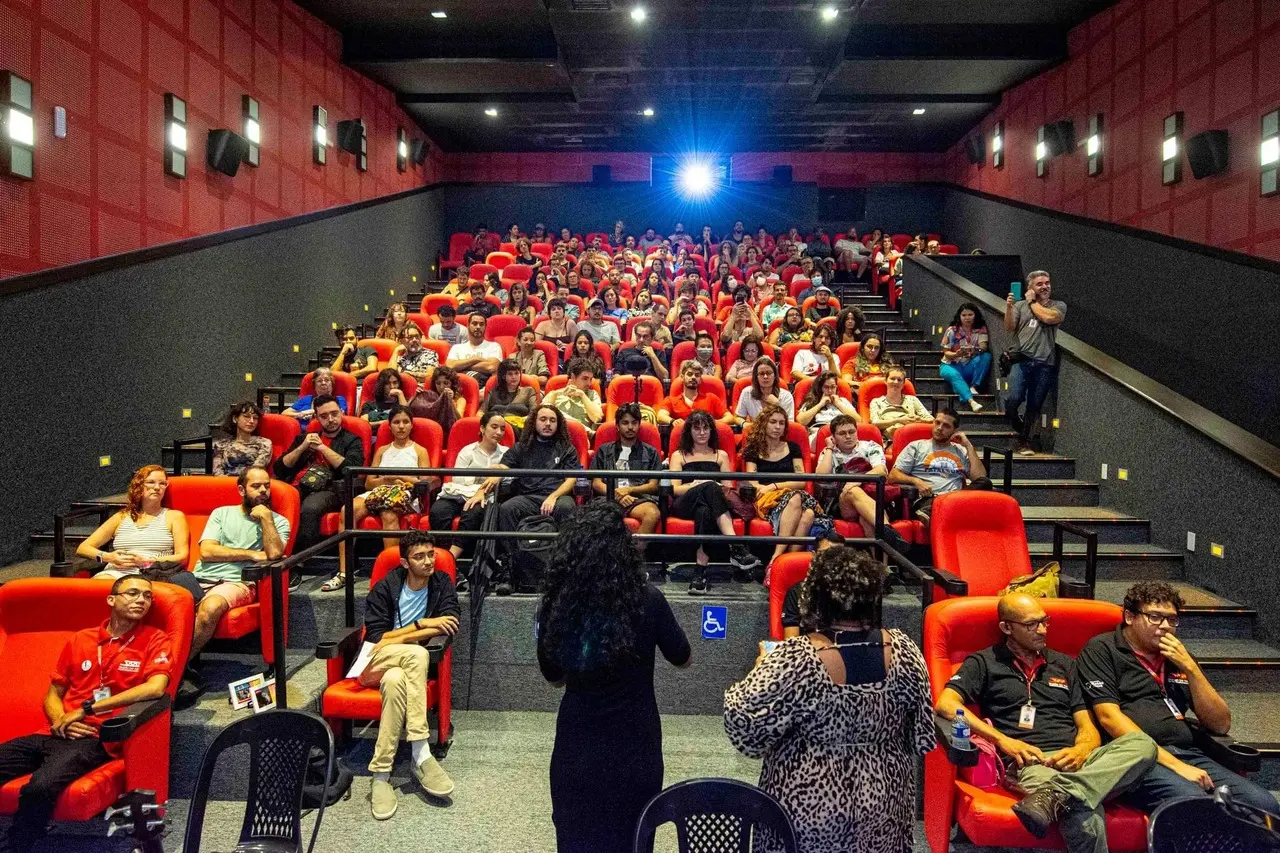 Uma plateia lota os assentos vermelhos de uma sala de cinema com paredes escuras e detalhes geométricos em vermelho nas laterais. Duas mulheres, de costas para a câmera na parte inferior central, parecem conduzir um debate ou apresentação para o público atento.
