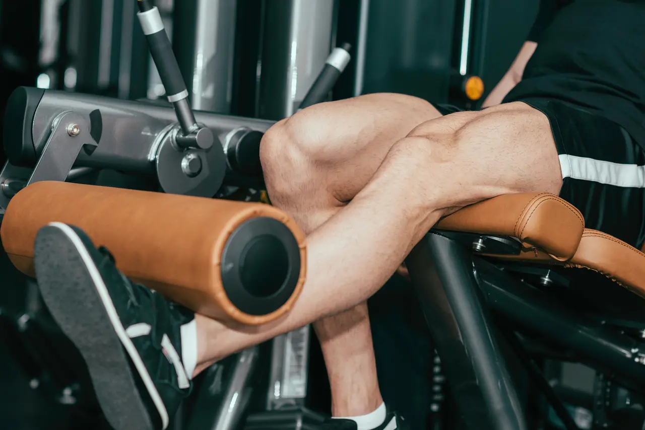 Imagem mostra um exercício em academia com homem apoiando os pés em uma máquina de extenção de perna, com as pernas estendendo-se sobre o equipamento enquanto trabalha resistência e força.