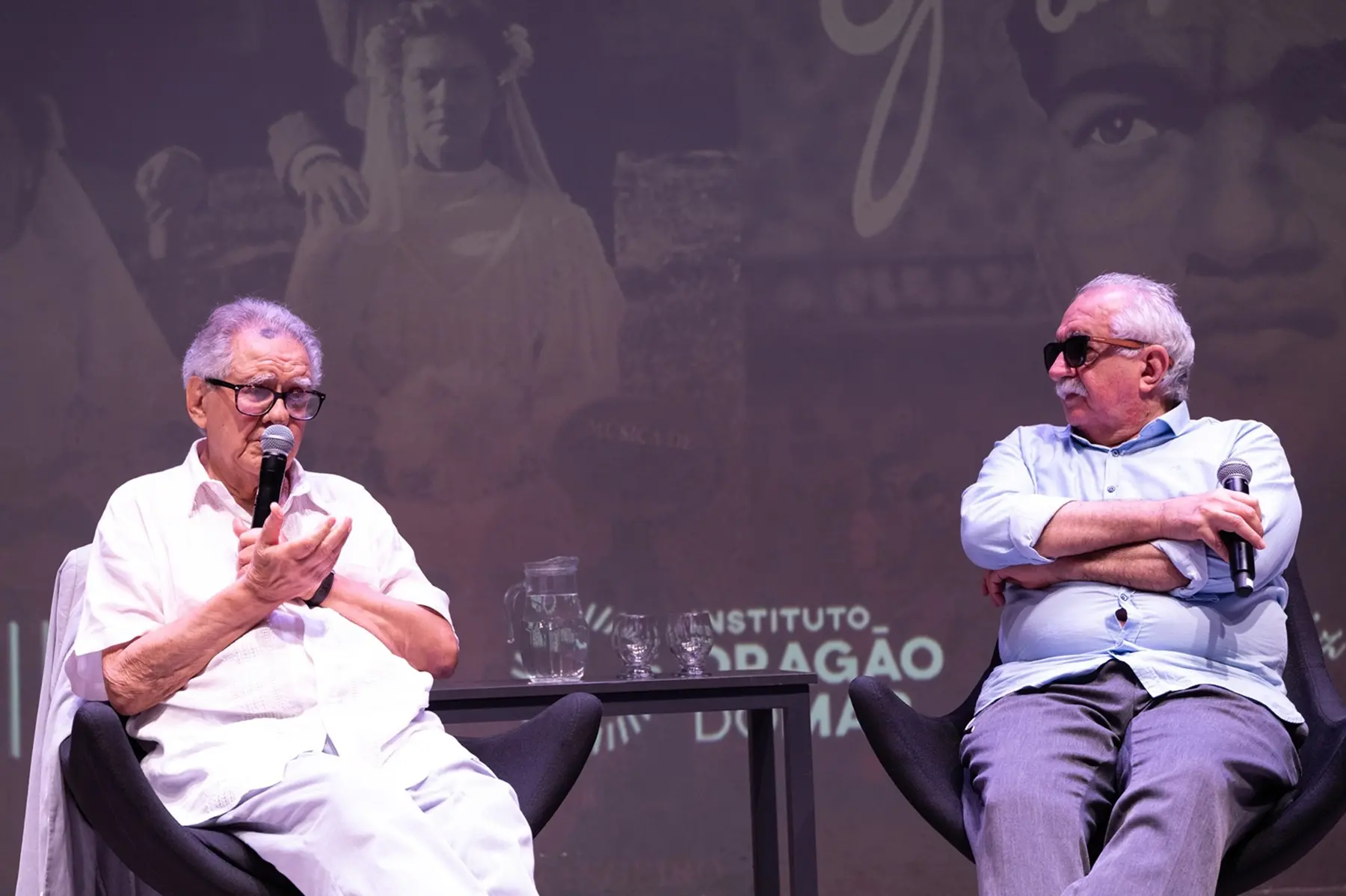 Os cineastas Luiz Carlos Barreto e Rosemberg Cariry estão sentados lado a lado em poltronas pretas durante um evento no palco. À esquerda, Luiz Carlos Barreto fala ao microfone vestindo roupa branca, enquanto Rosemberg Cariry, à direita, usa camisa azul, óculos escuros e mantém os braços cruzados segurando outro microfone.
