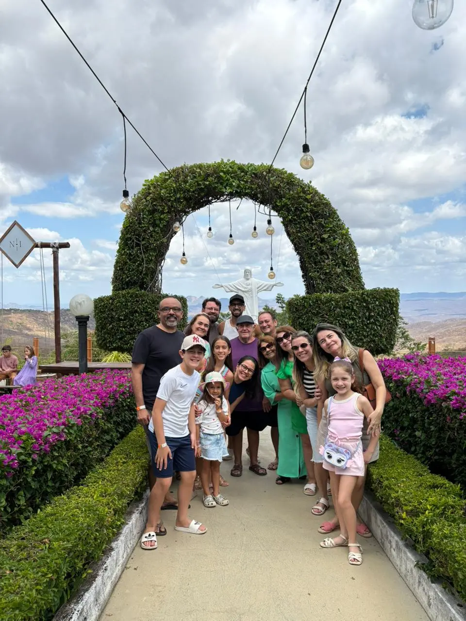 Na imagem, fotografia em um mirante ao ar livre durante o dia, sob um céu azul com nuvens brancas. No centro da imagem, cerca de 15 pessoas entre adultos e crianças estão reunidas e sorrindo para a foto. O grupo está posicionado em uma passarela de cimento com floreiras laterais repletas de flores cor-de-rosa (buganvílias). Acima do grupo, há um grande arco feito de vegetação verde decorado com várias lâmpadas penduradas por fios. Ao fundo, no centro do arco, vê-se uma pequena estátua branca do Cristo Redentor de braços abertos e, mais ao longe, uma paisagem montanhosa. A iluminação é natural e o clima é de confraternização.