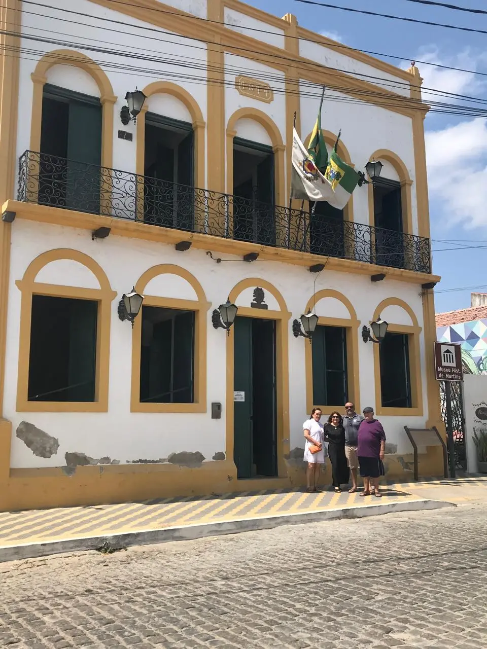 Na imagem, uma foto de ângulo médio capturando a fachada de dois andares de um edifício colonial histórico que funciona como o Museu Histórico de Martins. O prédio é pintado de branco com molduras amarelas brilhantes em torno de todas as portas e janelas. O primeiro andar tem um longo balcão de ferro forjado preto com cinco portas de sacada altas e em arco. O térreo tem quatro janelas em arco e uma porta principal central, todas com portas de madeira escura. Duas bandeiras (provavelmente do Brasil e do Rio Grande do Norte) estão hasteadas na sacada acima da porta principal. À direita da porta principal, no nível da calçada de paralelepípedos e calçamento de pedra, um grupo de quatro pessoas posa para a câmera: uma mulher de vestido branco, uma mulher de vestido preto, um homem de camisa de botões e bermuda e um homem de camisa roxa e bermuda escura. Uma placa de rua marrom e branca com o ícone do museu e o texto 