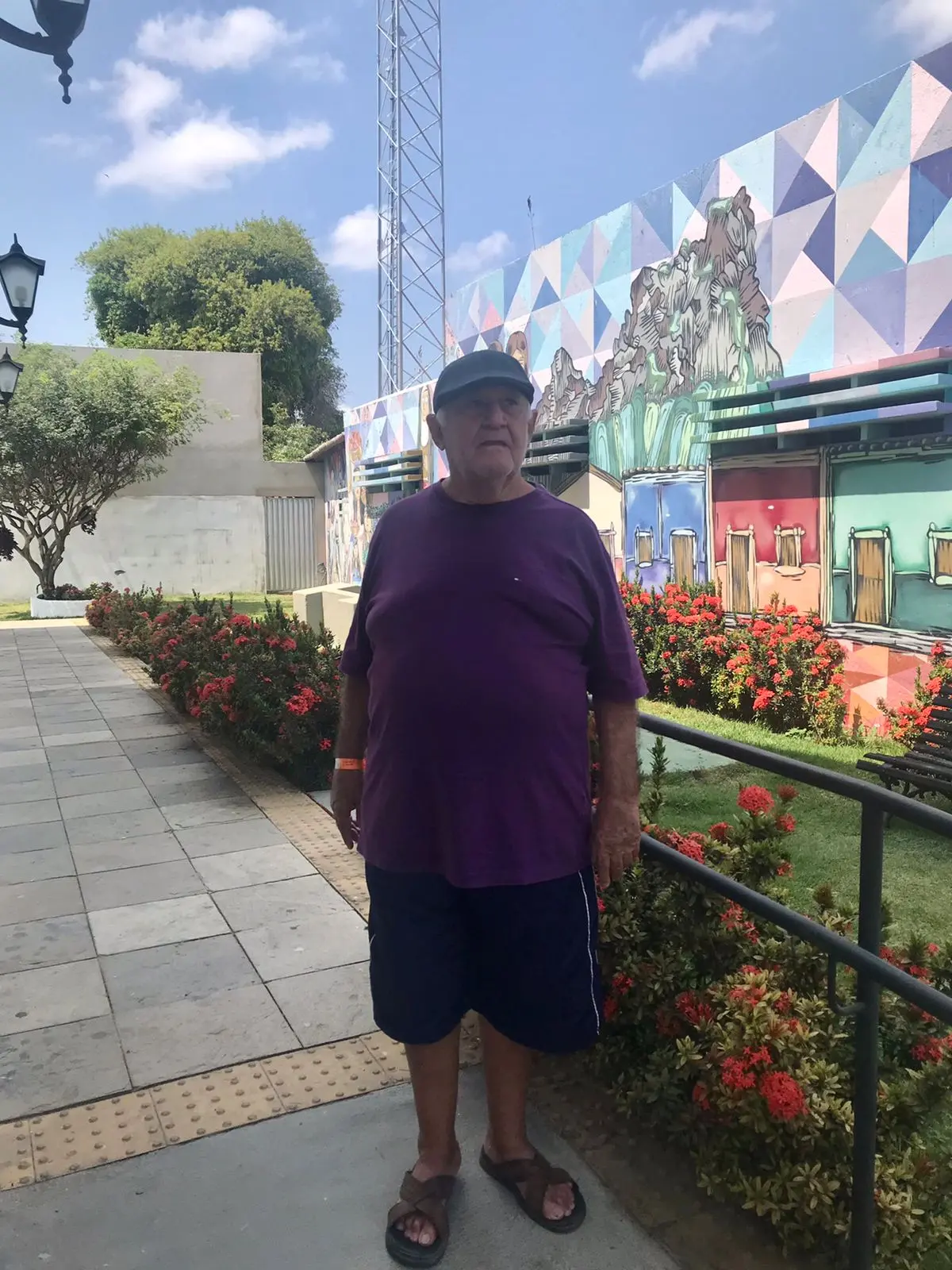 Na imagem, um homem idoso está em pé sobre uma calçada de blocos de concreto com piso tátil. Ele usa um boné preto, uma camiseta roxa larga, shorts esportivos azul-marinho e chinelos. Ele olha para o lado e para cima. Ao fundo, à direita, há um grande mural de arte urbana colorido em uma parede, com padrões geométricos e representações estilizadas de casas e montanhas. Canteiros com arbustos de flores vermelhas e uma grade de proteção preta flanqueiam o caminho. Ao fundo, à esquerda, há árvores, um prédio e postes de luz. O céu está azul claro com nuvens brancas esparsas.