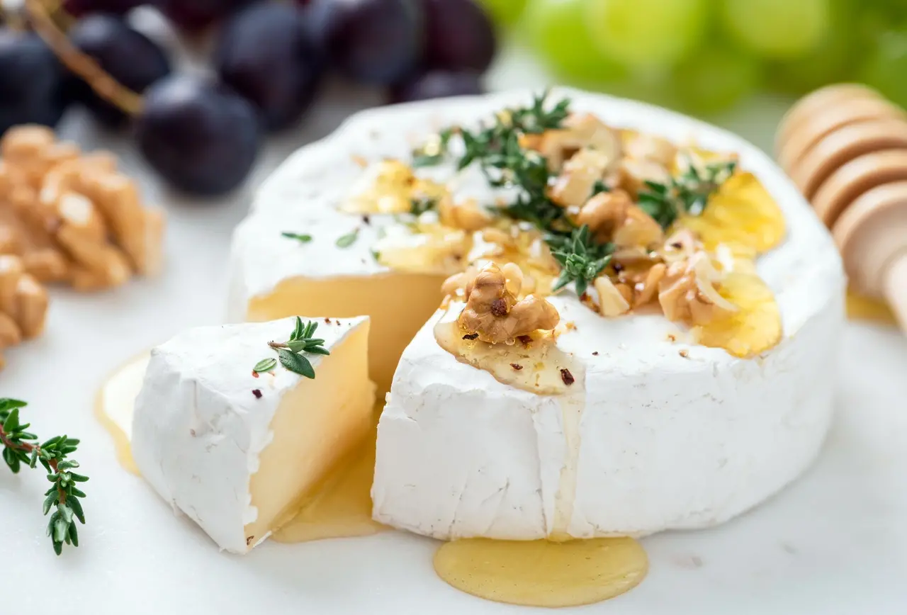 Queijo branco camembert, também conhecido como queijo brie, em pedaços sobre tábua, servido com mel e finalizado com ervas frescas, ao lado de uvas e nozes em fundo verde desfocado.