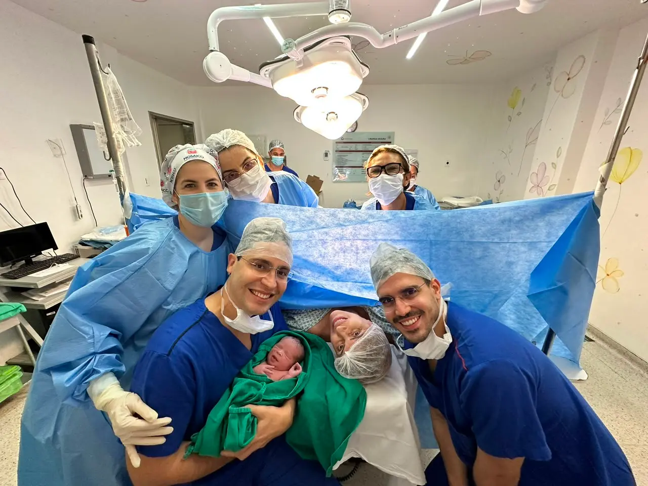 Na imagem, foto tirada no centro cirúrgico logo após um parto. Em primeiro plano, dois homens com roupas cirúrgicas azuis sorriem para a câmera; o da esquerda segura o bebê recém-nascido envolto em uma manta verde. Entre os dois homens, na mesa de cirurgia, a mãe sorri deitada usando touca descartável. Atrás de um lençol cirúrgico azul, a equipe médica composta por profissionais de saúde com aventais, toucas e máscaras cirúrgicas também posa sorrindo. O ambiente é iluminado por focos cirúrgicos no teto, e a parede ao fundo possui desenhos delicados de flores.