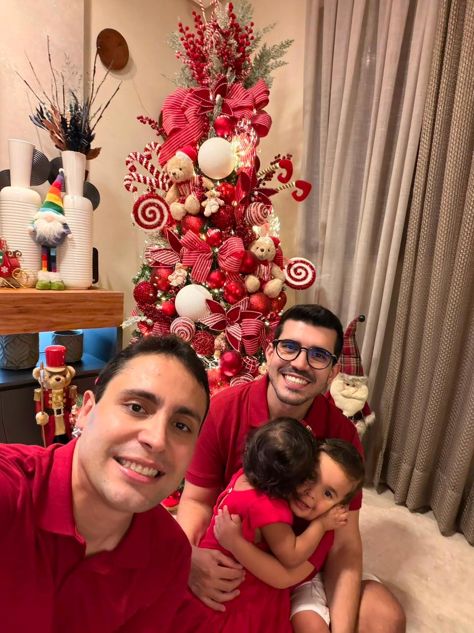 Na imagem, uma família de quatro pessoas tira uma foto sorridente em clima natalino. Em primeiro plano, dois homens vestem camisas polo vermelhas; o homem em segundo plano usa óculos e segura nos braços duas crianças pequenas, que também usam roupas vermelhas e se abraçam carinhosamente. Ao fundo, há uma árvore de Natal altamente decorada em tons de vermelho e branco, com laços grandes, ursinhos de pelúcia e bengalas de doces, além de cortinas beges e enfeites natalinos na sala.