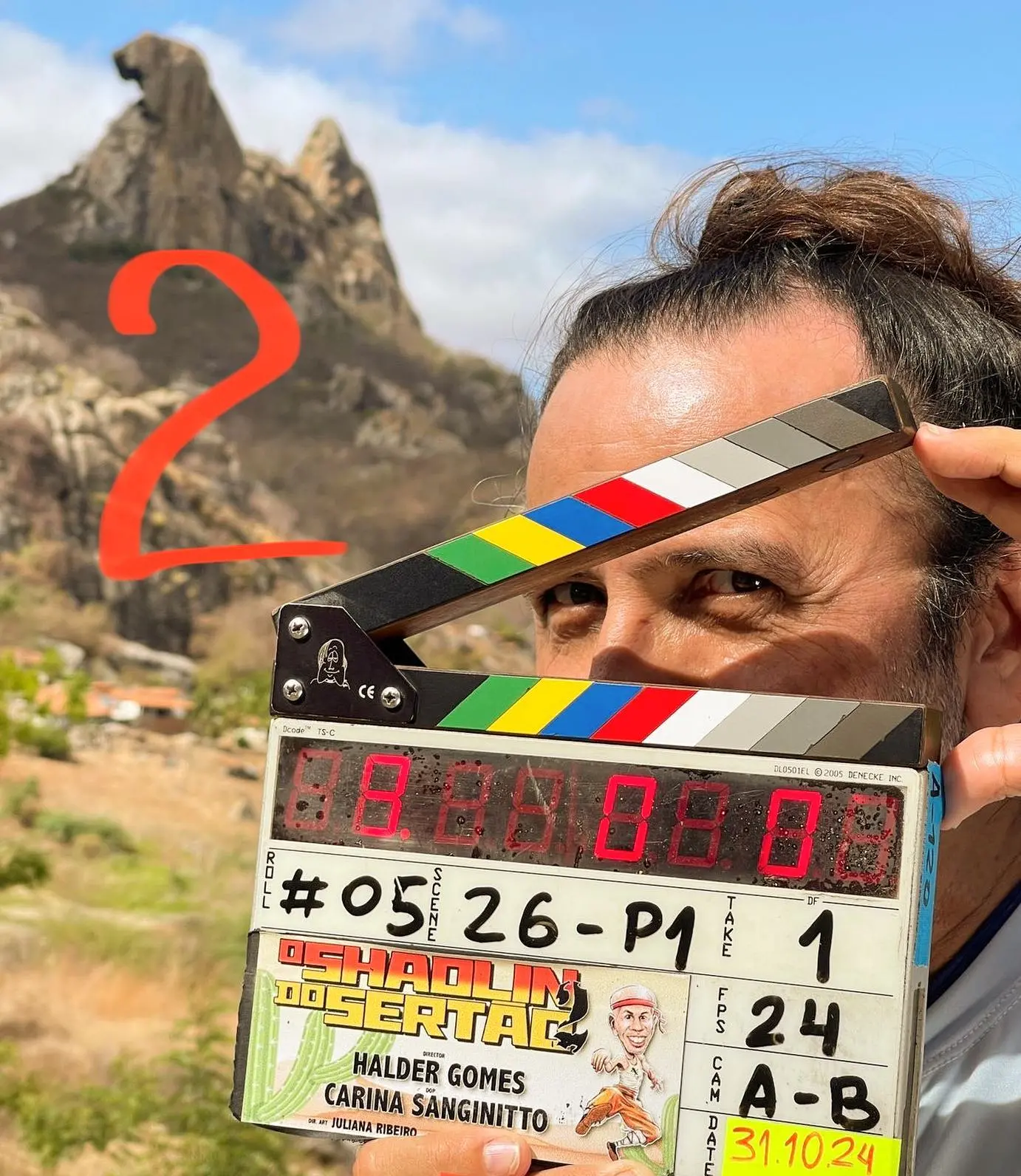 O cineasta Halder Gomes segura uma claquete de cinema com o título 'O Shaolin do Sertão 2' cobrindo parte do seu rosto. Ao fundo, sob o céu ensolarado, destaca-se uma formação rochosa da paisagem sertaneja com o número '2' desenhado em vermelho.