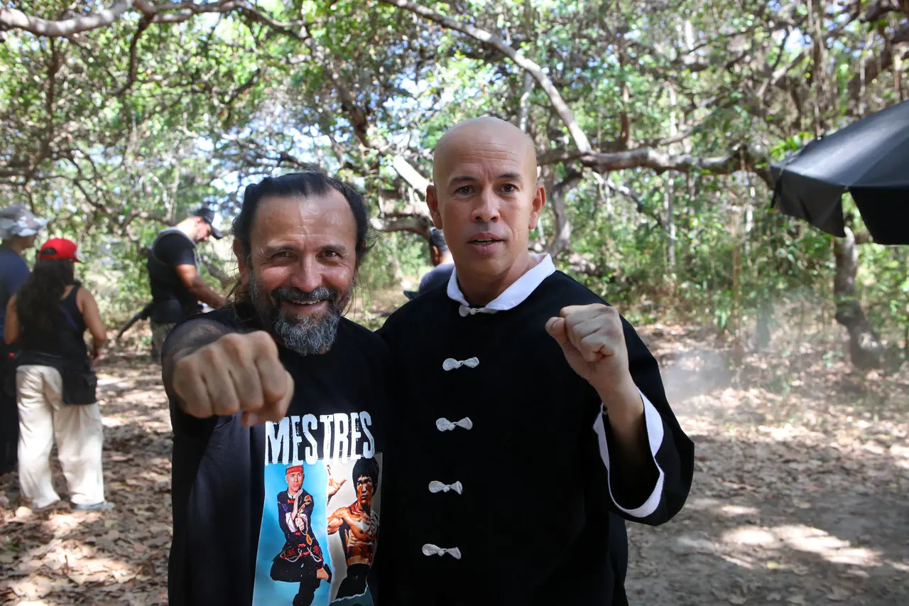 Halder Gomes e Edmilson Filho posam juntos ao ar livre em uma área arborizada, ambos fazendo pose com os punhos fechados em riste. Halder sorri usando camiseta preta estampa 'MESTRES', enquanto Edmilson veste uma túnica preta de artes marciais estilo Kung Fu.