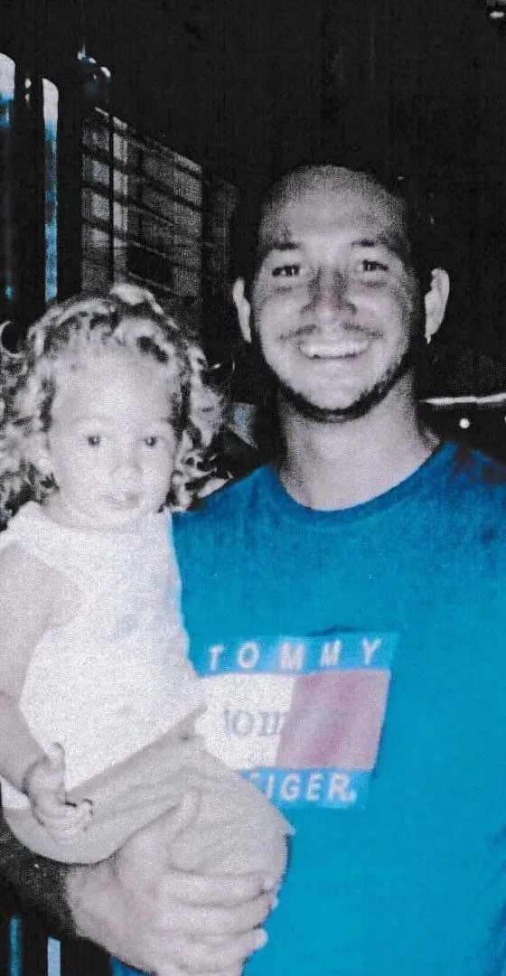 Na imagem, fotografia em plano médio mostrando um homem jovem sorrindo abertamente enquanto segura uma criança pequena no colo. O homem tem cabelos escuros presos para trás, barba rala e veste uma camiseta azul com a estampa do logotipo da Tommy Hilfiger no peito. A criança, à esquerda, é uma menina loira com cabelos cacheados e volumosos, vestindo uma regata clara. O fundo está escuro e desfocado, sugerindo um ambiente noturno ou interno com pouca iluminação.