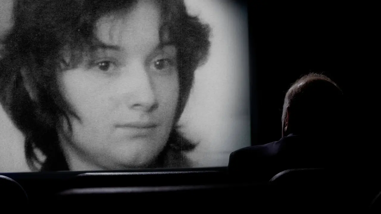 Um homem sentado em uma sala escura observa uma grande tela de cinema, vista de costas em silhueta. Na tela iluminada, é exibido o retrato em preto e branco de uma mulher jovem de cabelos curtos.