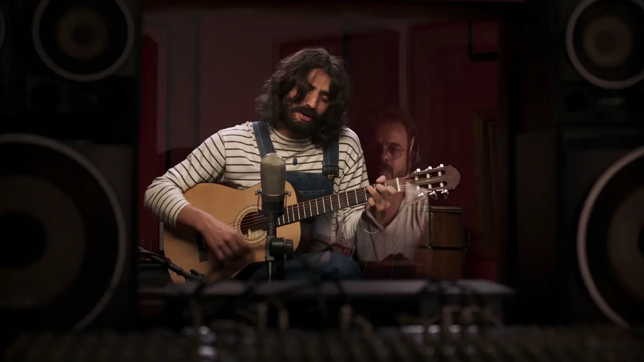O ator Caian Zattar interpreta Belchior e aparece como um homem de cabelos longos e barba que toca violão em um estúdio de gravação, vestindo uma blusa listrada e jardineira jeans diante de um microfone vintage. A cena é enquadrada por caixas de som e a mesa de som no primeiro plano, com o reflexo suave de outra pessoa no vidro ao fundo sob iluminação acolhedora.