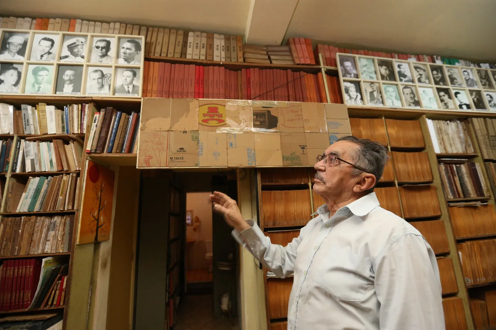 Na imagem, fotografia de baixo para cima de um homem idoso em perfil de três quartos, olhando para o alto com atenção. Ele usa camisa social clara de mangas compridas, tem cabelos curtos escuros e óculos de grau. O ambiente é um acervo ou biblioteca repleta de prateleiras de madeira altas cheias de livros, pastas e cadernos antigos. Na parte superior das prateleiras, há quadros emoldurados com retratos em preto e branco de diversas pessoas. Sobre a passagem de uma porta no centro, há uma estrutura adaptada feita de pedaços de papelão.