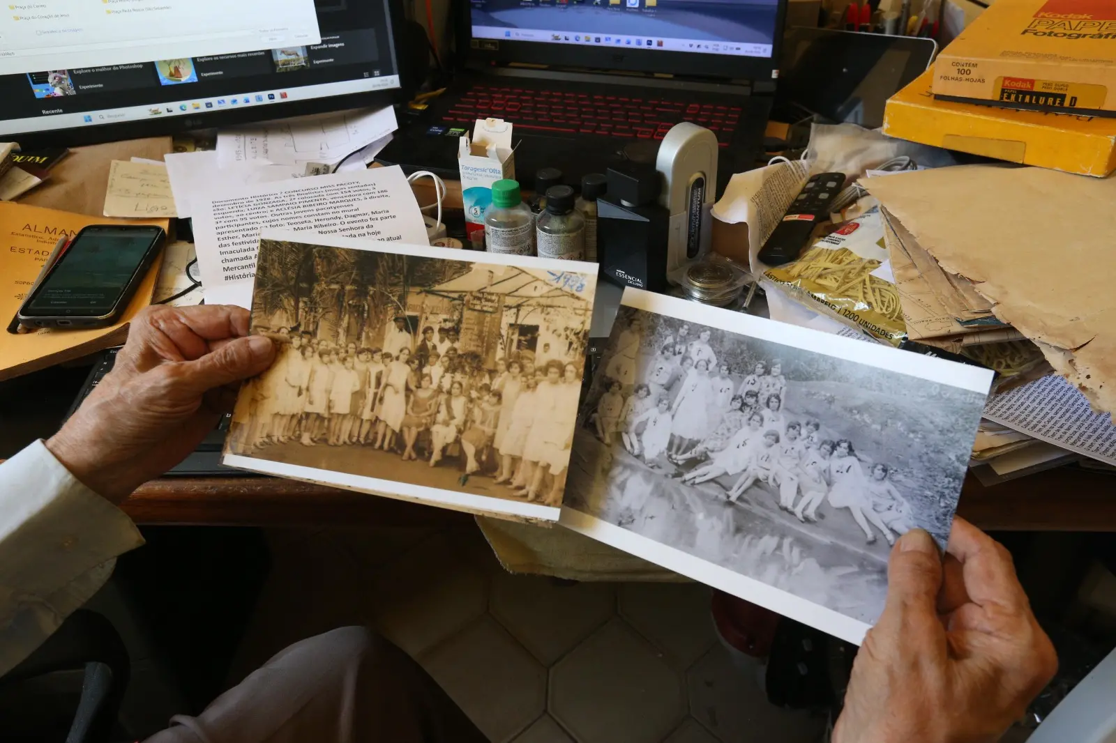Na imagem, fotografia em plano detalhe que mostra o ponto de vista de uma pessoa idosa segurando duas fotografias antigas em preto e branco sobre uma mesa de trabalho movimentada. À esquerda, a foto mostra um grande grupo de pessoas reunidas ao ar livre. À direita, a outra foto retrata um grupo de jovens e crianças sentados em um gramado. Ao fundo, a mesa está repleta de objetos, incluindo monitores de computador, um notebook aberto, papéis impressos, um smartphone, frascos de remédio e um perfume, caixas amarelas de papel fotográfico Kodak e papéis antigos acumulados.