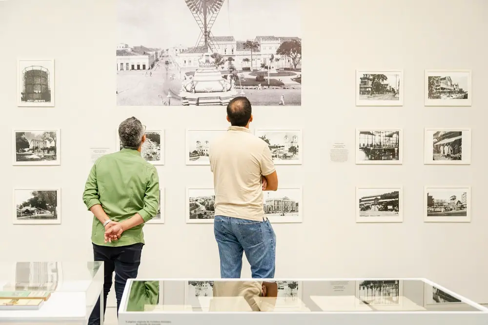 Na imagem, dois homens vistos de costas observam uma parede branca repleta de fotografias em preto e branco com molduras brancas em uma exposição de arte. O homem à esquerda veste camisa verde de mangas compridas dobradas até os cotovelos e calça escura, com as mãos cruzadas atrás das costas. O homem à direita usa camisa polo bege e calça jeans. Ao centro e acima deles, destaca-se uma fotografia ampliada do centro histórico de uma cidade com um grande monumento. Em primeiro plano, na parte inferior, há vitrines transparentes exibindo documentos e livros históricos.