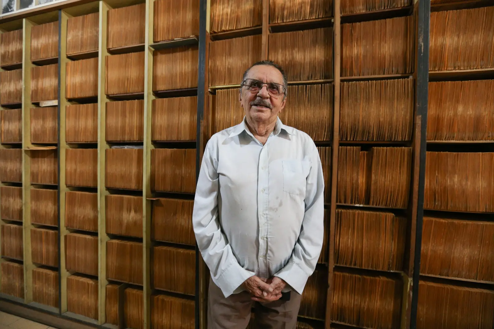 Na imagem, um homem idoso de óculos e bigode está em pé no centro da imagem, sorrindo suavemente com as mãos cruzadas à frente do corpo. Ele veste uma camisa social clara de mangas compridas e calça marrom. Ao fundo, há grandes estantes de madeira repletas de prateleiras organizadas e completamente lotadas de discos de vinil antigos em capas beges e amareladas.