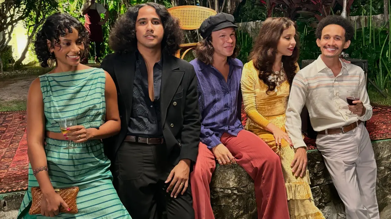 Parte do elenco da série 'Alucinação' vestindo figurinos em estilo vintage dos anos 1970 posa junta ao ar livre em um ambiente arborizado. O grupo interage sorridente e descontraído, posando sobre tapetes e uma mureta de pedra, com destaque para a composição vibrante de cores e texturas das roupas.