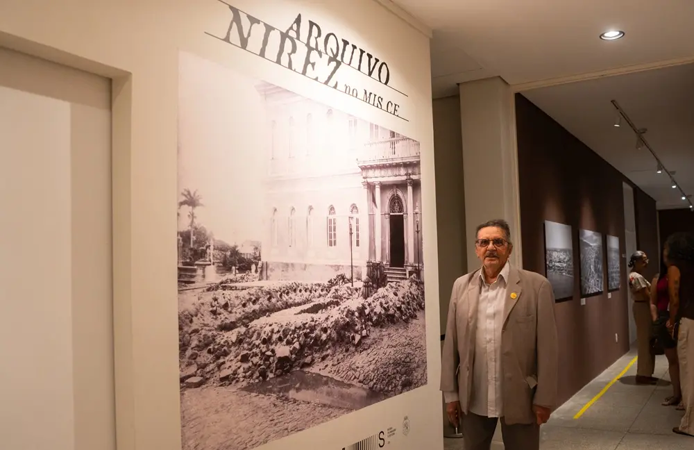 Na imagem, um homem idoso de terno bege e óculos está em pé ao lado de um painel de uma exposição. O painel traz uma grande fotografia histórica em tom sépia de um edifício antigo e a inscrição 