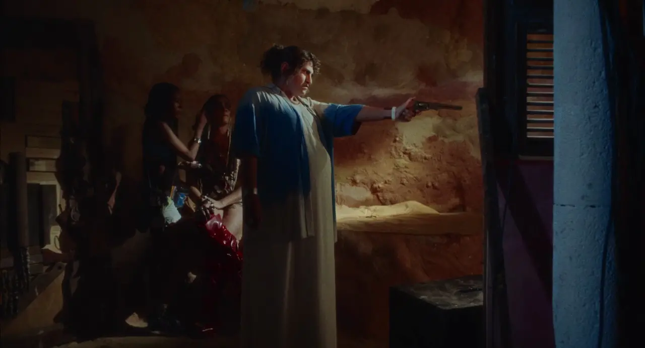 Cena da atriz Noá Bonoba no filme cearense 'Morte e Vida Madalena' mostra a personagem grávida, vestindo uma túnica clara com camisão azul e apontando um revólver para frente em um ambiente rústico iluminado. Ao fundo, à esquerda, duas figuras observam a cena na penumbra num cenário com paredes de tom terra e iluminação dramática.