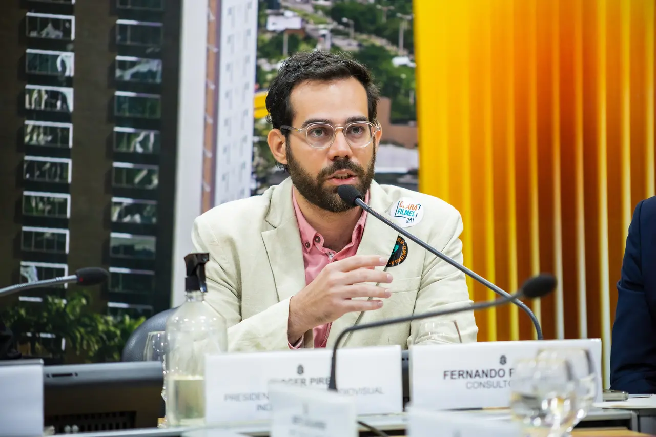 Produtor Roger Pires, de óculos e barba, participa de uma reunião em mesa de conferência, falando ao microfone. À frente há placas de identificação, copos e uma garrafa. Ao fundo, aparecem prédios desfocados e painéis verticais em tons de amarelo e laranja. Um adesivo com o texto 'Ceará Filmes Já!' está fixado na lapela do blazer claro.