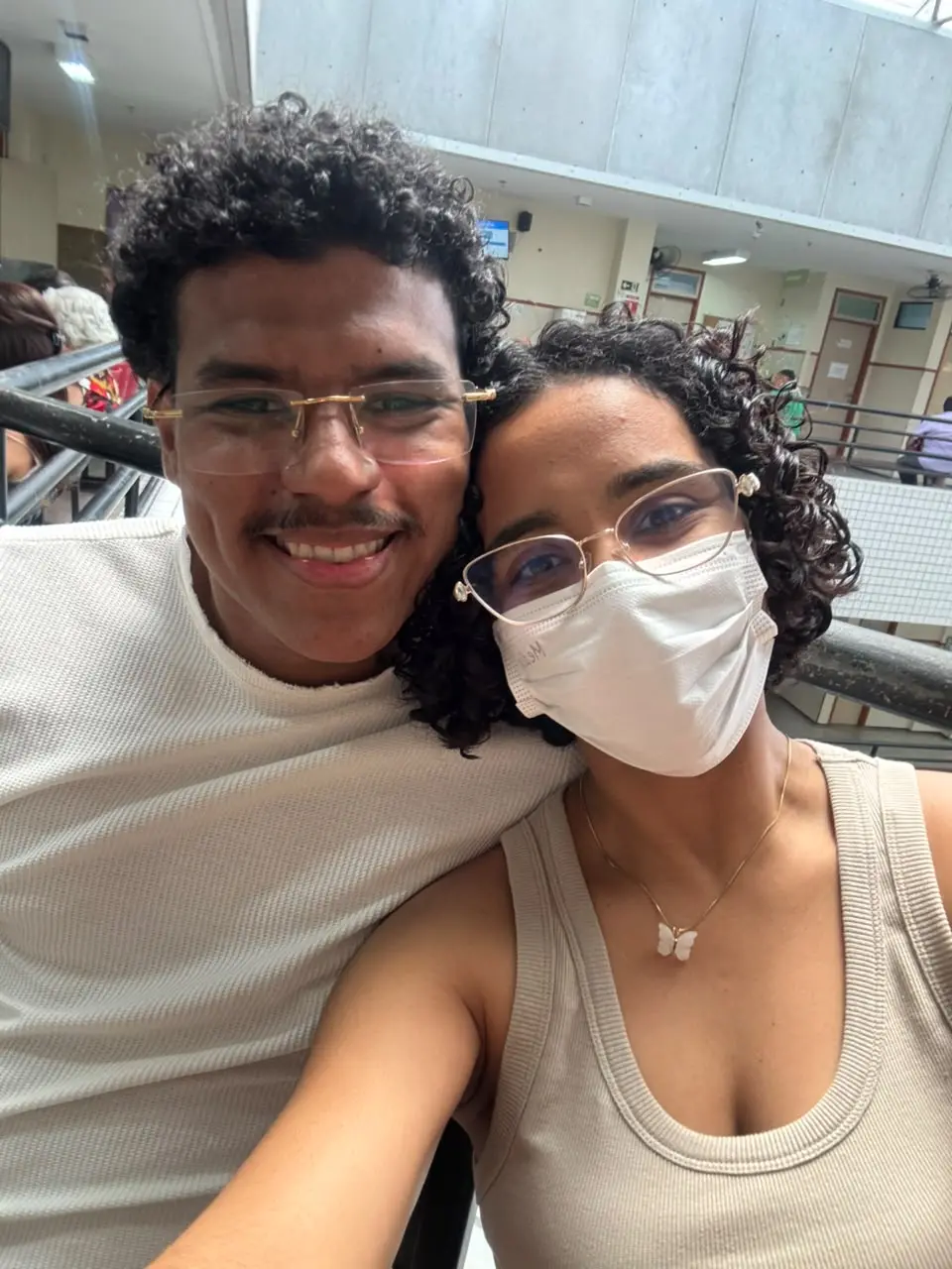 Na imagem, selfie de duas pessoas sentadas lado a lado em um ambiente interno com corredores, guarda-corpos e circulação de pessoas ao fundo. Ambas usam óculos. À esquerda, uma pessoa veste camiseta branca de manga curta. À direita, outra pessoa veste blusa regata clara, usa máscara facial branca e colar com pingente em formato de borboleta. A fotografia foi tirada em plano próximo, destacando os rostos e a parte superior do corpo, com iluminação natural vindo da área superior do ambiente.