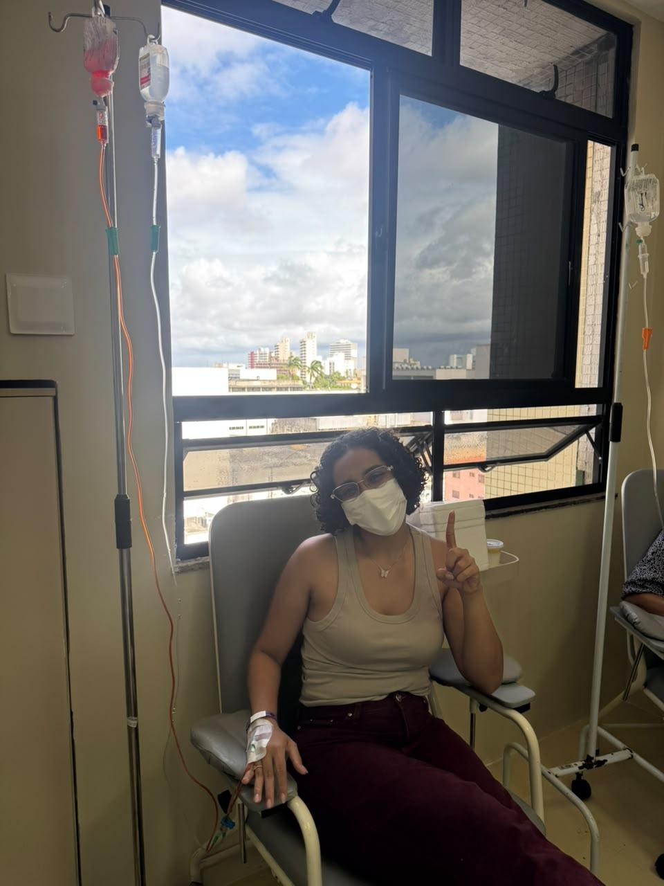 Na imagem, pessoa sentada em uma poltrona de atendimento em ambiente hospitalar ou clínica, usando máscara facial e óculos. Um acesso venoso está fixado na mão esquerda, conectado a equipamentos de infusão ao lado da cadeira. A pessoa levanta um dedo indicador enquanto está posicionada diante de uma grande janela com vista para edifícios e céu nublado. À esquerda e à direita aparecem suportes com bolsas de soro e outros materiais utilizados em procedimentos médicos. O ambiente é iluminado por luz natural vinda da janela.