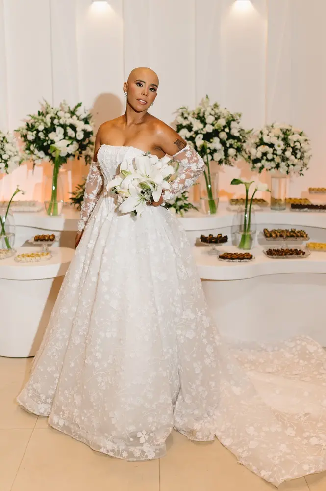 Na imagem, pessoa posa em pé usando vestido de casamento branco com detalhes florais bordados, mangas rendadas transparentes e longa cauda estendida sobre o piso. A pessoa segura um buquê de lírios brancos à frente do corpo. Ao fundo, há uma mesa de recepção com arranjos florais brancos em vasos de vidro e bandejas de doces distribuídas sobre superfícies brancas curvas. O ambiente interno é iluminado por luz suave e decoração em tons claros.