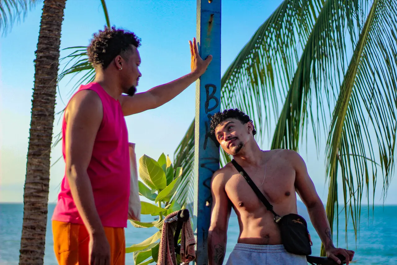Duas pessoas estão próximas a um poste azul em uma área à beira-mar. À esquerda, uma pessoa veste regata rosa e bermuda laranja, apoiando uma das mãos no poste. À direita, outra pessoa está sem camisa, usa bermuda clara e bolsa transversal preta, apoiada no mesmo poste. Ao fundo aparecem coqueiros, vegetação, o mar e o céu azul iluminado pela luz do dia.