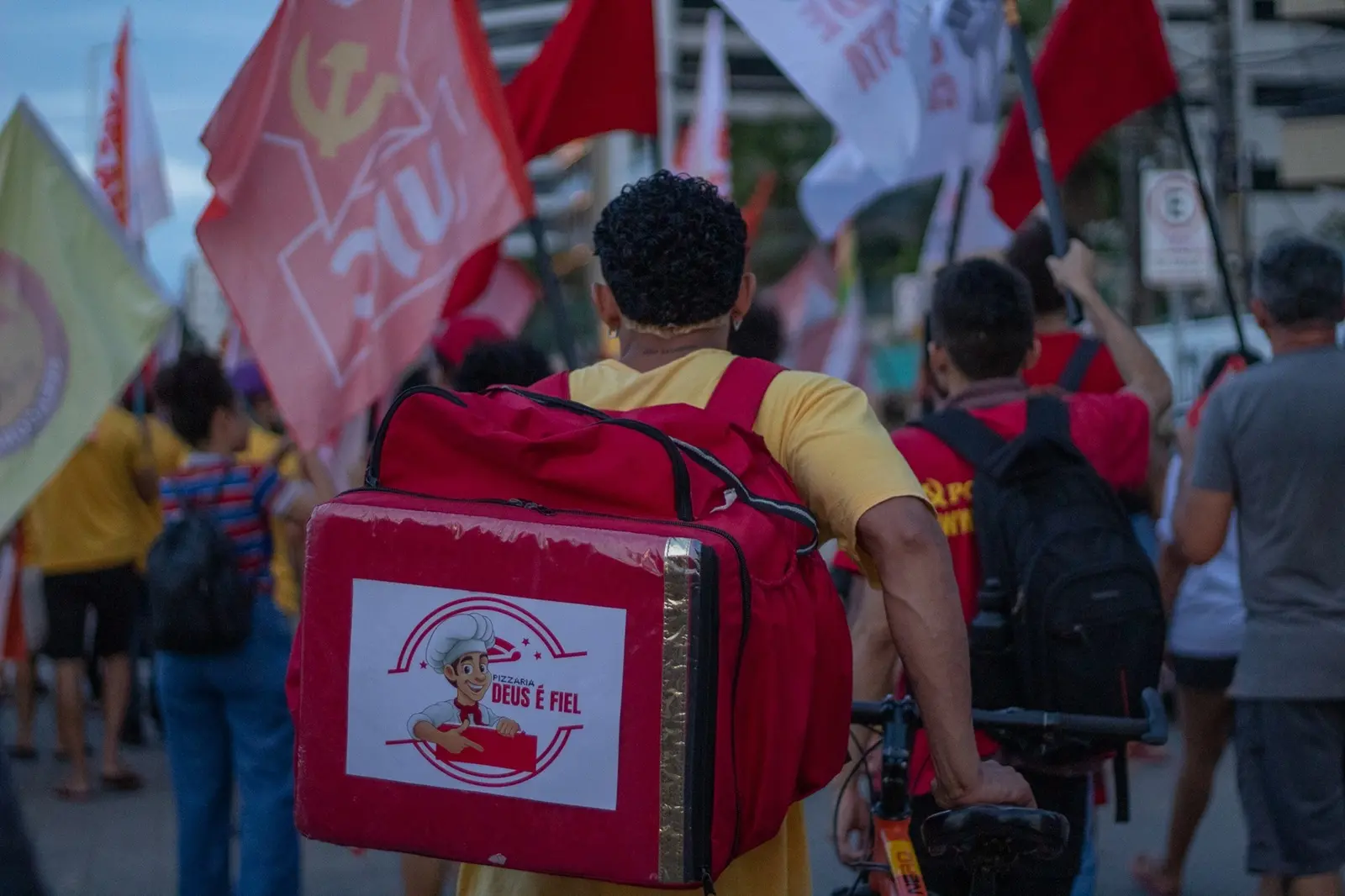 Pessoa conduz uma bicicleta em meio a uma manifestação de rua, carregando uma grande mochila vermelha de entregas com o logotipo de uma pizzaria. Ao redor, várias pessoas caminham segurando bandeiras vermelhas e brancas, em um ambiente urbano ao entardecer.