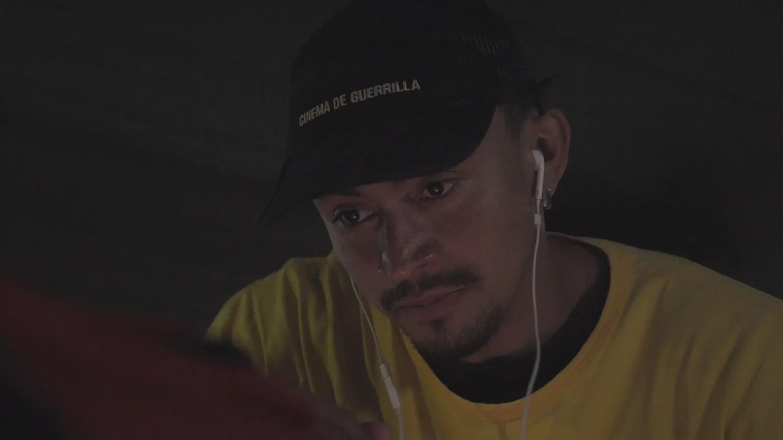 Pessoa usando camiseta amarela, boné preto com a inscrição 'Cinema de Guerrilla' e fones de ouvido observa uma tela fora do enquadramento em ambiente escuro. A iluminação suave destaca o rosto e os fios dos fones, enquanto o fundo permanece desfocado e com pouca luz.