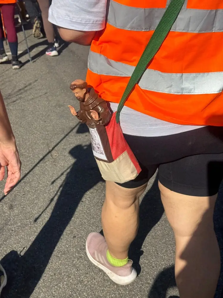 Na imagem, pessoa vista de costas caminha por uma via asfaltada durante um evento ou peregrinação ao ar livre. A pessoa veste colete laranja com faixas refletivas, camiseta branca, bermuda escura e tênis claros. A tiracolo, carrega uma bolsa de tecido com uma pequena imagem religiosa presa ao lado externo. Ao redor, aparecem outras pessoas caminhando na mesma direção. A luz do sol projeta sombras alongadas sobre o asfalto, evidenciando a atividade realizada em espaço aberto.
