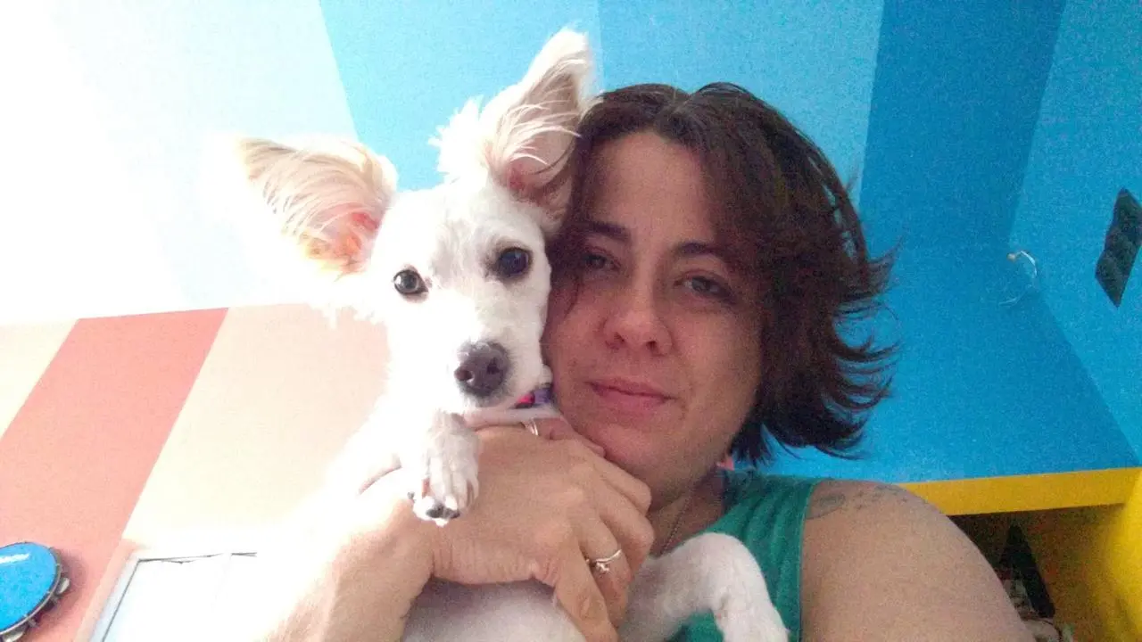 Na imagem, pessoa segura um cachorro pequeno de pelagem branca em um ambiente interno com paredes coloridas em tons de azul. O animal está apoiado sobre o braço da pessoa e tem orelhas grandes e eretas. A pessoa veste uma regata verde-clara e aparece em primeiro plano ao lado do cachorro. Ao fundo, há uma parede azul, uma porta e uma área iluminada pela luz natural, que destaca o pelo claro do animal.