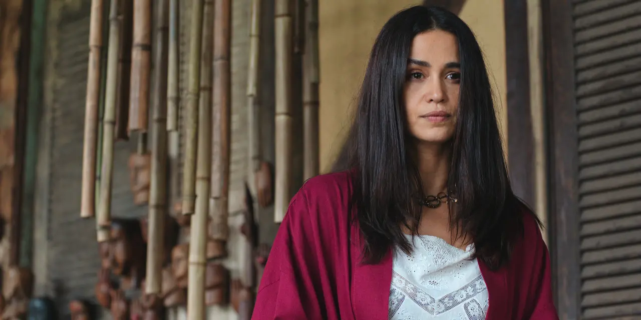 Pessoa de cabelos longos e escuros veste blusa branca e cardigan vinho, em pé diante de uma parede com elementos decorativos de bambu e madeira. A imagem tem enquadramento fechado e fundo desfocado.